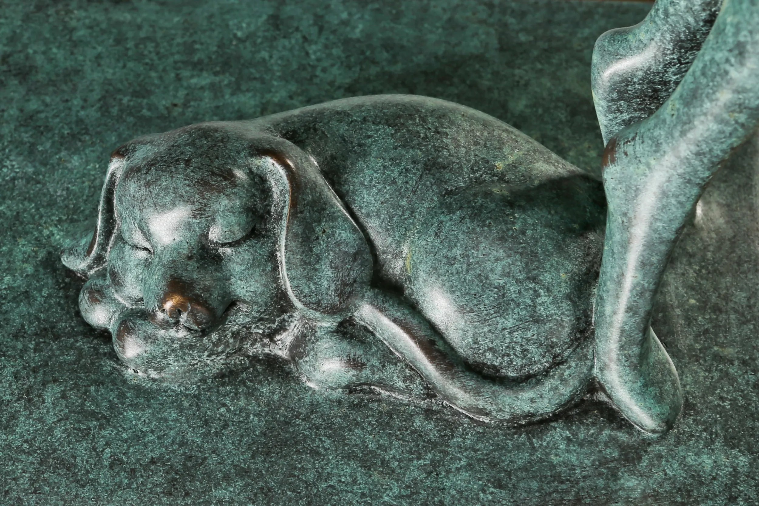 Close-up of the sleeping bronze dog beside the woman's feet with green patina and bronze highlights