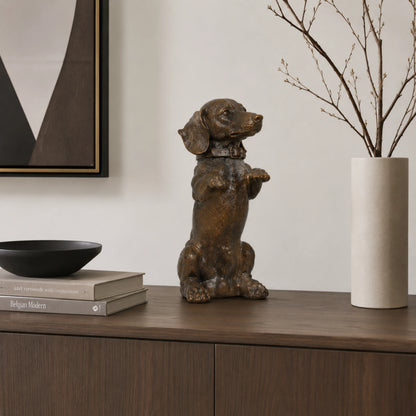 A charming bronze dachshund statue in a playful upright seated pose with front paws raised begging, highlighting its long body and short legs.