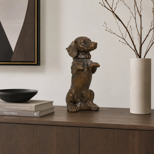 A charming bronze dachshund statue in a playful upright seated pose with front paws raised begging, highlighting its long body and short legs.