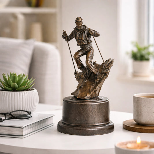 This is a dynamic bronze sculpture of a skier, crafted using the lost-wax casting method. The piece captures the athlete mid-action, navigating a rugged, snow-like terrain with ski poles firmly in hand and legs bent in a powerful stance. The rich, warm bronze patina accentuates the fluid folds of the skier’s jacket and pants, as well as the textured, naturalistic base that simulates a snow-covered slope.
