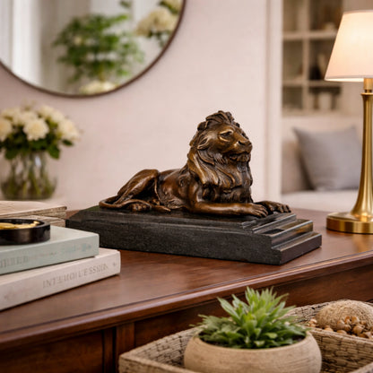 This is a majestic bronze lion sculpture, cast using the lost-wax method, depicting the animal in a poised, reclining stance. The sculpture features a rich, warm bronze patina with a smooth, polished finish that accentuates the lion’s powerful musculature and the intricate, flowing details of its mane. Its head is held high, conveying a sense of quiet authority and regal composure, while its body rests in a relaxed yet alert posture.
