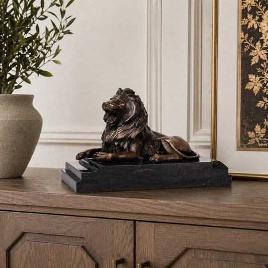 This is a powerful bronze lion sculpture, cast using the lost-wax method, depicting the animal in a reclining pose with its mouth open in a fierce roar. The sculpture features a rich, aged bronze patina that highlights the intricate, flowing details of its mane and the taut musculature of its body, conveying a sense of primal strength and authority. The lion’s head is raised, and its expression is intense, while its body rests in a relaxed yet alert posture, suggesting a moment of warning or dominance.
